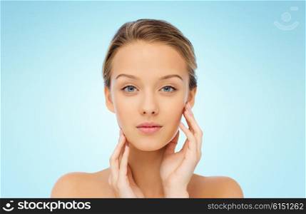 beauty, people and health concept - young woman with bare shoulders touching her face over blue background