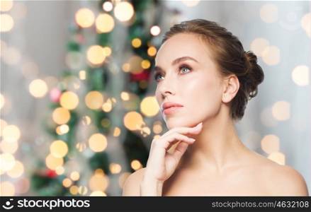 beauty, people and holidays concept - beautiful young woman touching her face over christmas tree lights background
