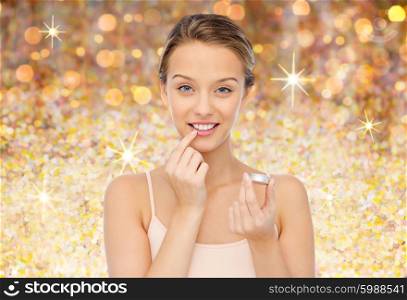 beauty, people and lip care concept - smiling young woman applying lip balm to her lips over golden glitter background