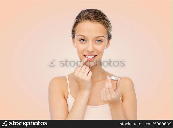 beauty, people and lip care concept - smiling young woman applying lip balm to her lips over beige background