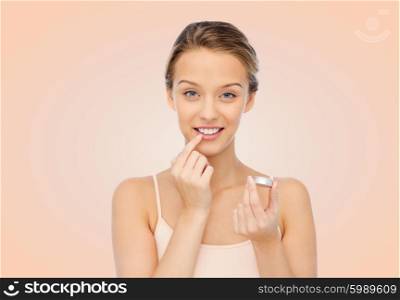 beauty, people and lip care concept - smiling young woman applying lip balm to her lips over beige background