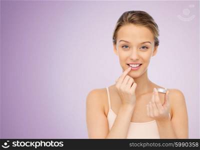 beauty, people and lip care concept - smiling young woman applying lip balm to her lips over violet background