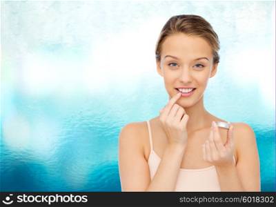 beauty, people and lip care concept - smiling young woman applying lip balm to her lips over blue water background