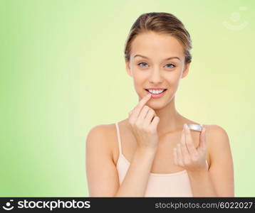 beauty, people and lip care concept - smiling young woman applying lip balm to her lips over green natural background