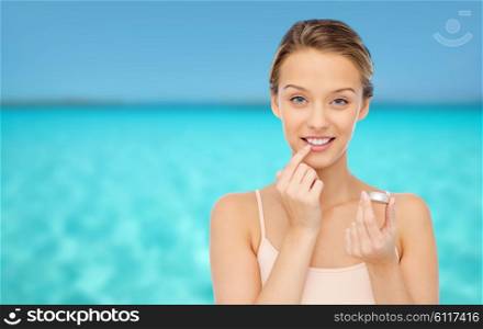 beauty, people and lip care concept - smiling young woman applying lip balm to her lips over blue sea and sky background