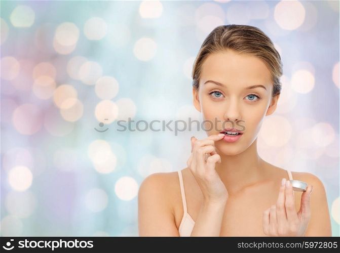 beauty, people and lip care concept - young woman applying lip balm to her lips over blue holidays lights background