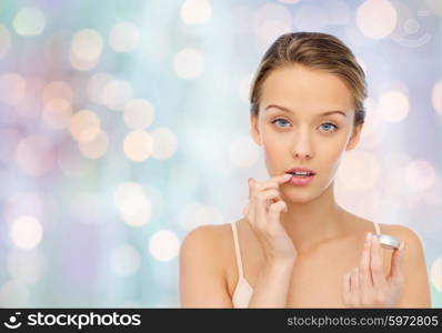 beauty, people and lip care concept - young woman applying lip balm to her lips over blue holidays lights background