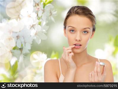 beauty, people and lip care concept - young woman applying lip balm to her lips over green natural background with cherry blossoms