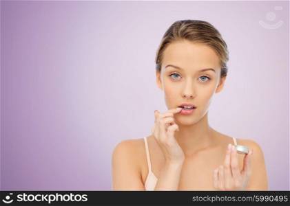 beauty, people and lip care concept - young woman applying lip balm to her lips over violet background