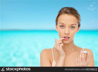 beauty, people and lip care concept - young woman applying lip balm to her lips over blue sea and sky background