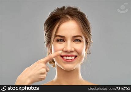beauty, people and rhinoplasty concept - beautiful young woman pointing to her nose over grey background. beautiful young woman pointing to her nose