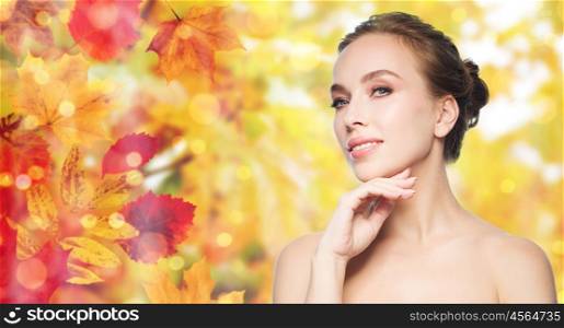 beauty, people and skin care concept - beautiful young woman touching her face over natural autumn leaves and lights background