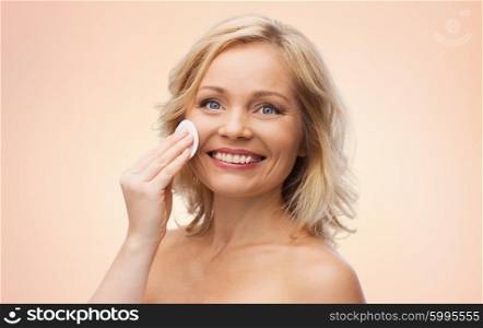 beauty, people and skincare concept - happy middle aged woman cleaning face and removing make up with cotton pad over beige background
