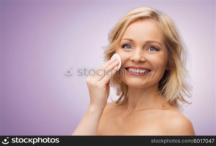 beauty, people and skincare concept - happy middle aged woman cleaning face and removing make up with cotton pad over violet background