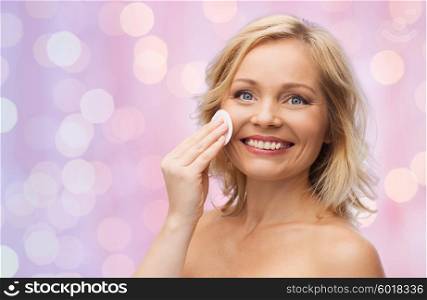 beauty, people and skincare concept - happy middle aged woman cleaning face and removing make up with cotton pad over pink holidays lights background