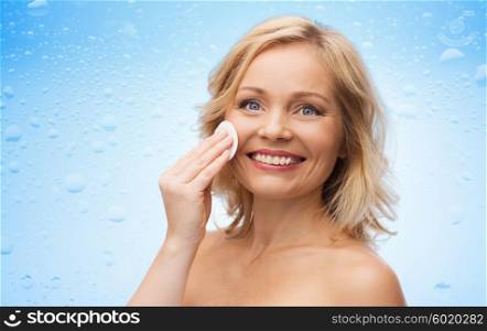 beauty, people and skincare concept - happy middle aged woman cleaning face and removing make up with cotton pad over water drops on blue background