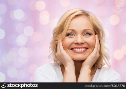 beauty, people and skincare concept - smiling middle aged woman in white shirt touching face over pink holidays lights background