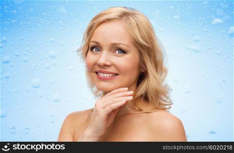 beauty, people and skincare concept - smiling middle aged woman with bare shoulders touching face over water drops on blue background