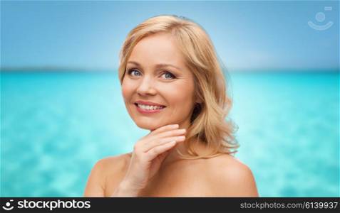 beauty, people and skincare concept - smiling middle aged woman with bare shoulders touching face over blue sea and sky background