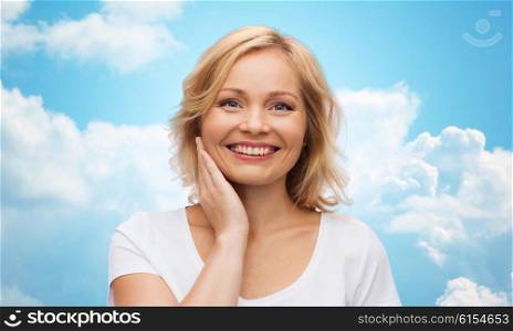 beauty, people and skincare concept - smiling woman in white shirt touching face over blue sky and clouds background