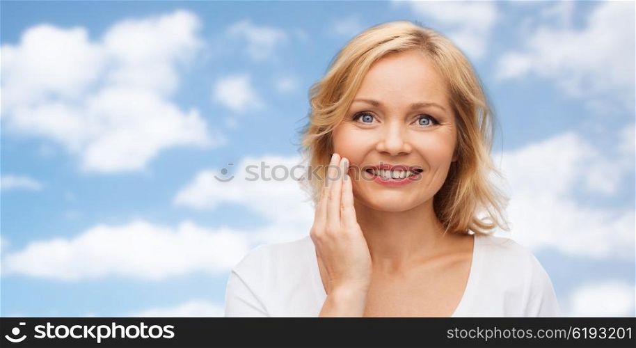 beauty, people and skincare concept - smiling woman in white shirt touching face over blue sky and clouds background