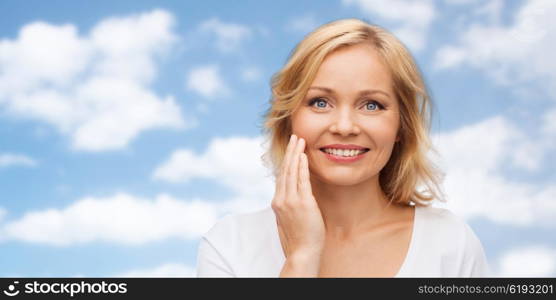 beauty, people and skincare concept - smiling woman in white shirt touching face over blue sky and clouds background