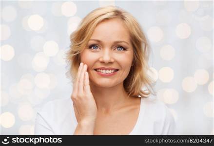 beauty, people and skincare concept - smiling woman in white shirt touching face over holidays lights background