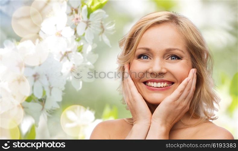 beauty, people and skincare concept - smiling woman with bare shoulders touching face over natural spring cherry blossom background