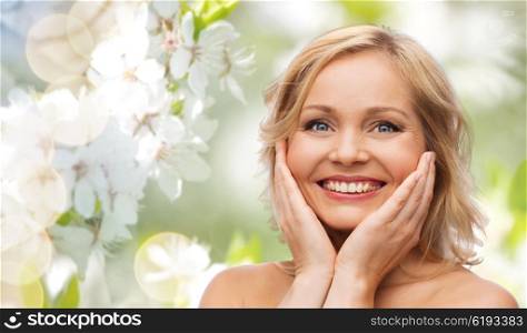 beauty, people and skincare concept - smiling woman with bare shoulders touching face over natural spring cherry blossom background
