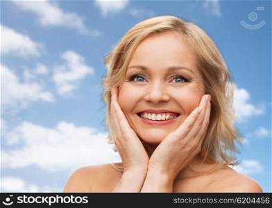 beauty, people and skincare concept - smiling woman with bare shoulders touching face over blue sky and clouds background