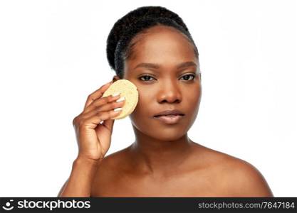 beauty, people and skincare concept - young african american woman with bare shoulders cleaning face with exfoliating sponge over white background. young woman cleaning face with exfoliating sponge