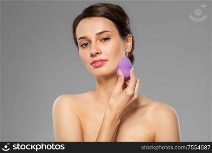 beauty, people and skincare concept - young woman applying foundation with make up blending sponge over grey background. young woman with sponge applying makeup