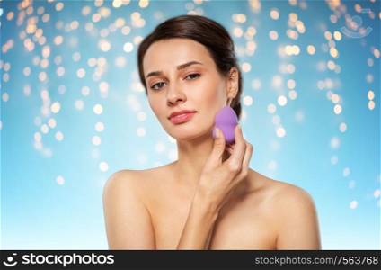 beauty, people and skincare concept - young woman applying foundation with make up blending sponge over holidays lights on blue background. young woman with sponge applying makeup