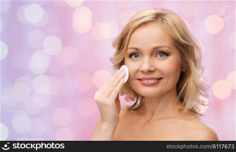 beauty, people and skincare concept - young woman cleaning face and removing make up with cotton pad over pink lights background