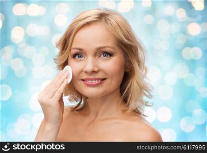 beauty, people and skincare concept - young woman cleaning face and removing make up with cotton pad over blue holidays lights background