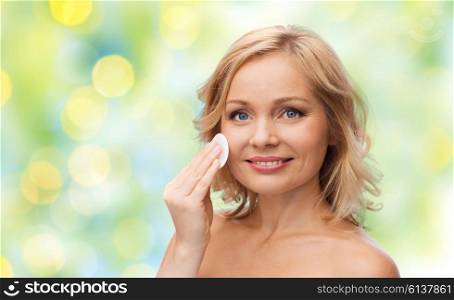 beauty, people and skincare concept - young woman cleaning face and removing make up with cotton pad over green summer lights background