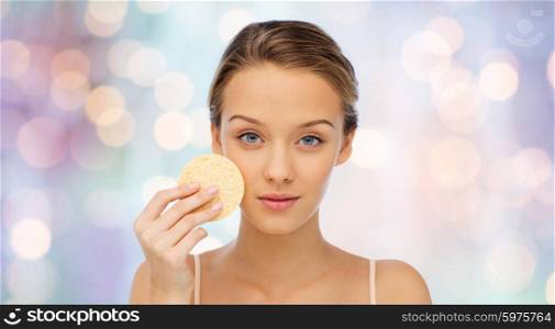 beauty, people and skincare concept - young woman cleaning face with exfoliating sponge over purple holidays lights background