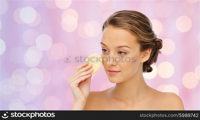 beauty, people and skincare concept - young woman cleaning face with exfoliating sponge over pink holidays lights background