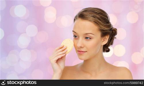 beauty, people and skincare concept - young woman cleaning face with exfoliating sponge over pink holidays lights background