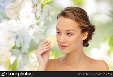 beauty, people and skincare concept - young woman cleaning face with exfoliating sponge over green natural cherry blossom background