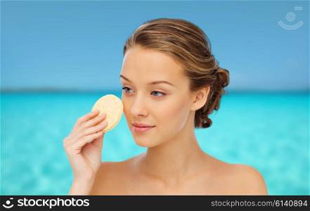 beauty, people and skincare concept - young woman cleaning face with exfoliating sponge over blue sea and sky background