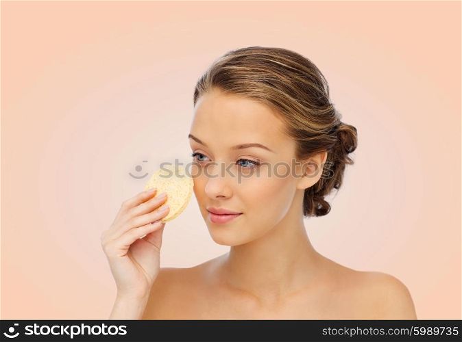beauty, people and skincare concept - young woman cleaning face with exfoliating sponge over beige background