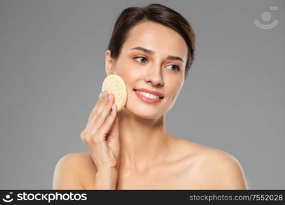 beauty, people and skincare concept - young woman cleaning face with exfoliating sponge over grey background. young woman cleaning face with exfoliating sponge