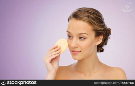 beauty, people and skincare concept - young woman cleaning face with exfoliating sponge over violet background