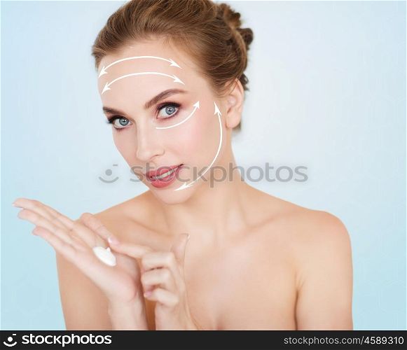 beauty, people, anti-age and cosmetics concept - happy young woman with moisturizing cream on hand and face lifting arrows over blue background