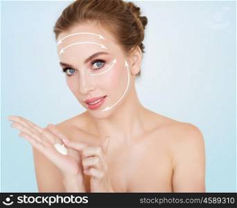 beauty, people, anti-age and cosmetics concept - happy young woman with moisturizing cream on hand and face lifting arrows over blue background