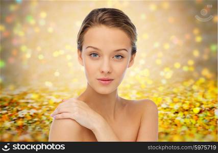 beauty, people, body care and health concept - smiling young woman face and hand on bare shoulder over golden holidays lights or yellow glitter background