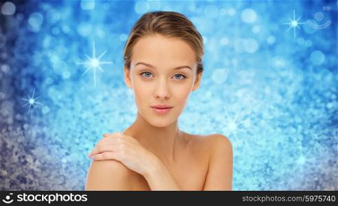 beauty, people, body care and health concept - smiling young woman face and hand on bare shoulder over blue holidays lights or glitter background