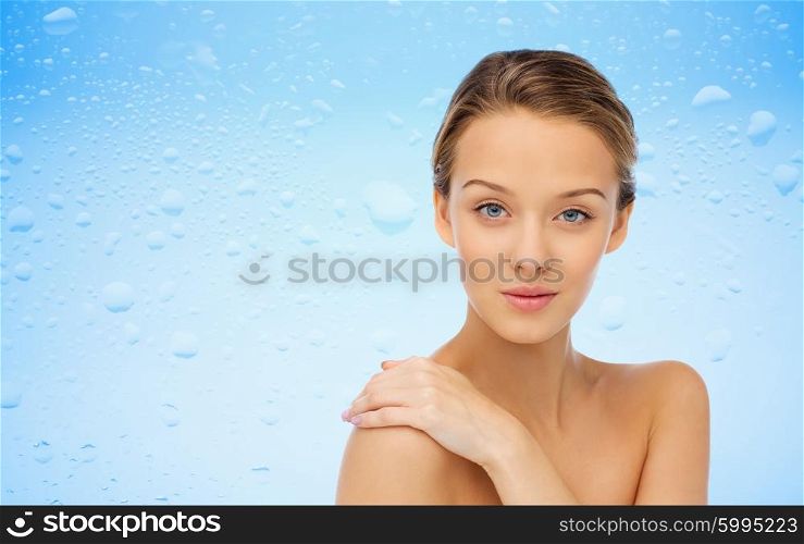 beauty, people, body care and health concept - smiling young woman face and hand on bare shoulder over water drops on blue background