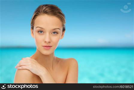 beauty, people, body care and health concept - smiling young woman face and hand on bare shoulder over blue sea and sky background
